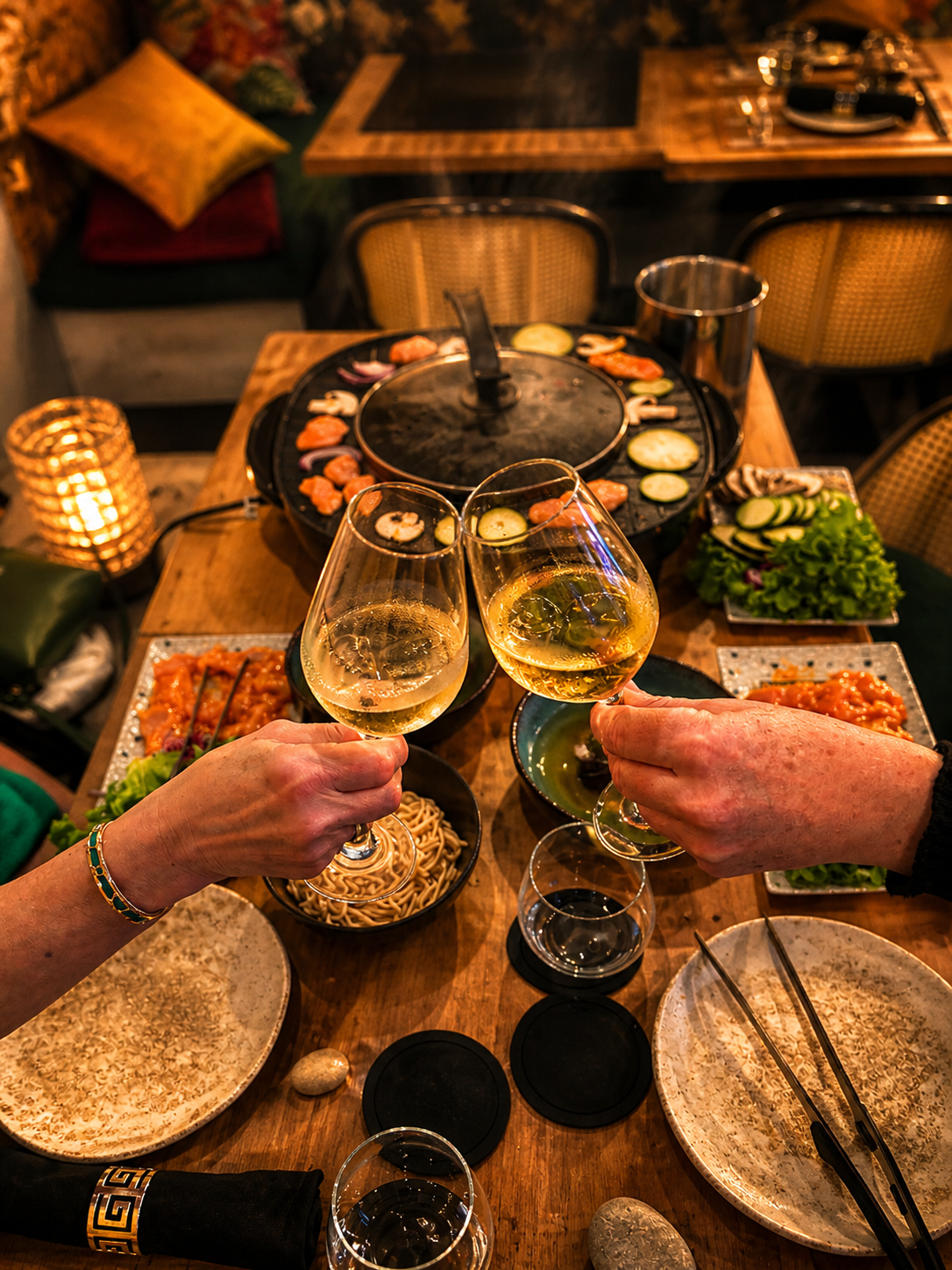 Deux convives trinquent au-dessus du grill coréen de leur table, entourés de viandes, de légumes et de nouilles